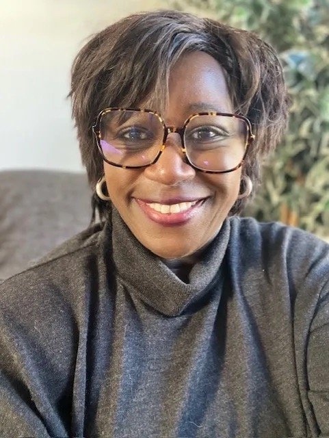 A smiling woman wearing glasses and a grey jumper, photographed in a relaxed indoor setting.